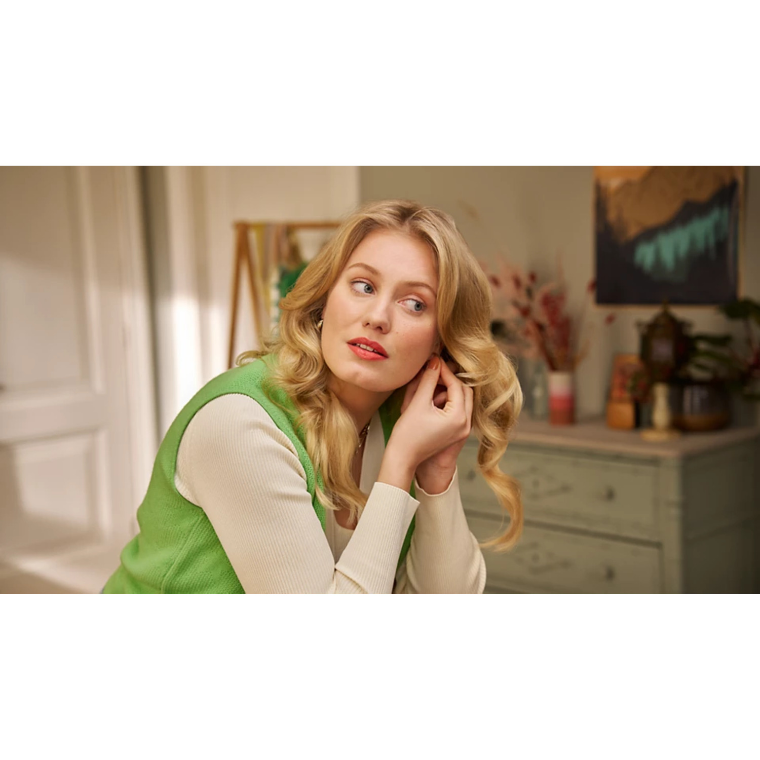 Blonde woman adjusting earring, soft focus background of a room with artwork and floral arrangements.