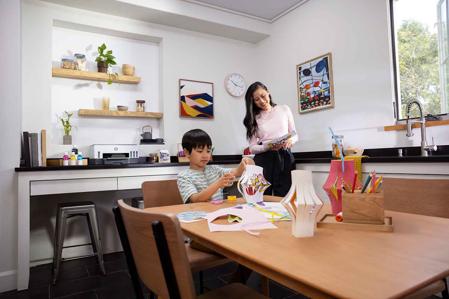 Een vrouw en een kind die knutselen aan een tafel met een HP-printer, gekleurd papier en knutselspullen.