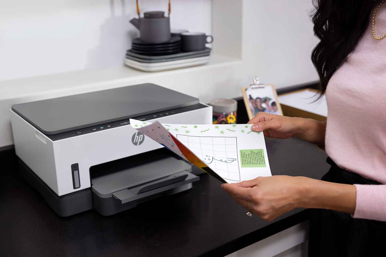 Een vrouw houdt een kleurrijk papier vast bij een wit-grijze printer, op een zwart aanrecht.