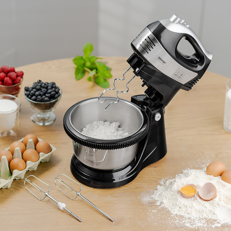 Mikser stojący ze składnikami na stole: jajkami, jagodami i mąką.