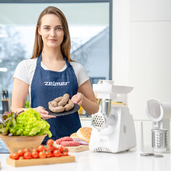 Kobieta w fartuchu trzymająca talerz z kiełbasami, z jedzeniem w tle.