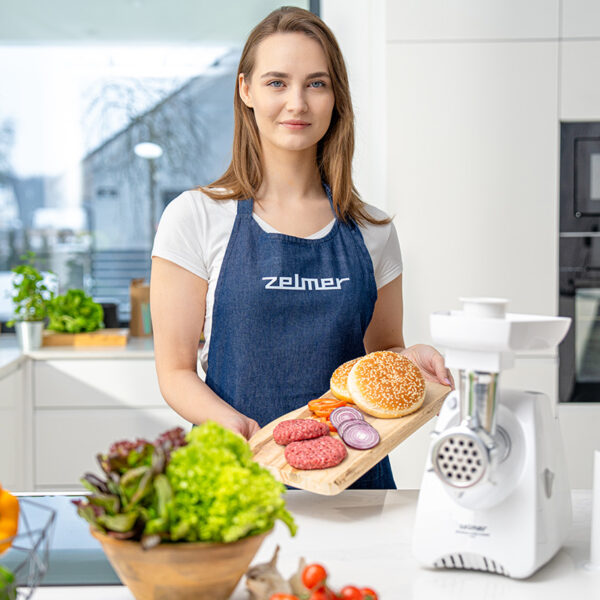 Kobieta w fartuchu trzyma deskę do krojenia z burgerami, warzywami i maszynką do mięsa.