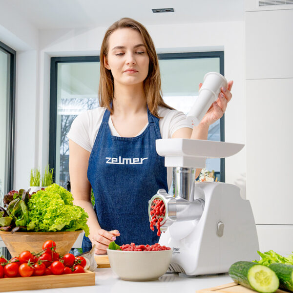Kobieta w fartuchu, dodająca składniki do białej maszynki do mięsa. Widoczne warzywa i miska mięsa.