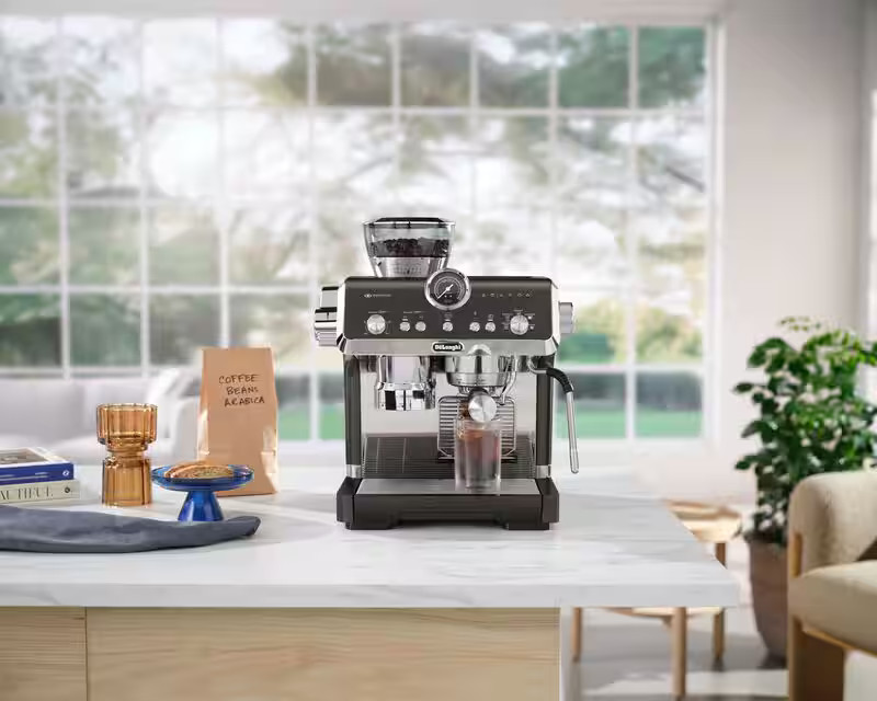 Ekspres do kawy na marmurowym blacie. Widoczna filiżanka, worek z ziarnami i rośliny.