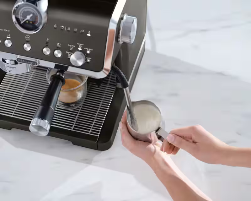 Ekspres do kawy, czarny i stalowy, z filiżanką kawy i dzbankiem do spieniania mleka.