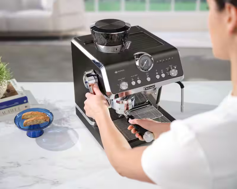 Kobieta obsługuje czarno-srebrny ekspres do espresso De'Longhi na białym stole.