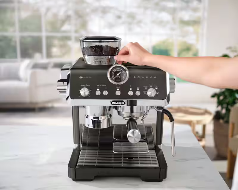 Ręka osoby reguluje pokrętło w ekspresie do kawy. Ziarna w przezroczystym pojemniku są na górze.