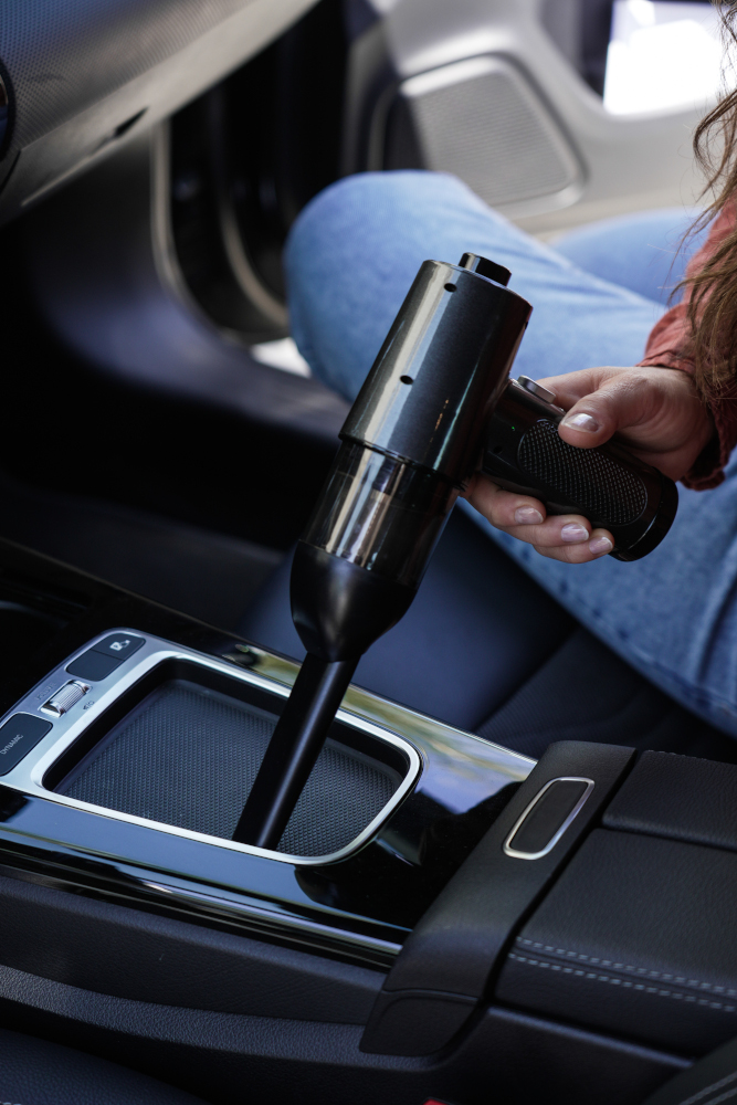 A person using a black hand vacuum inside a car. The vacuum is pointed towards a console.