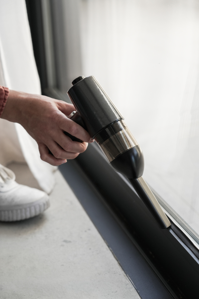 Hand holding a black and silver handheld vacuum cleaner near a window.