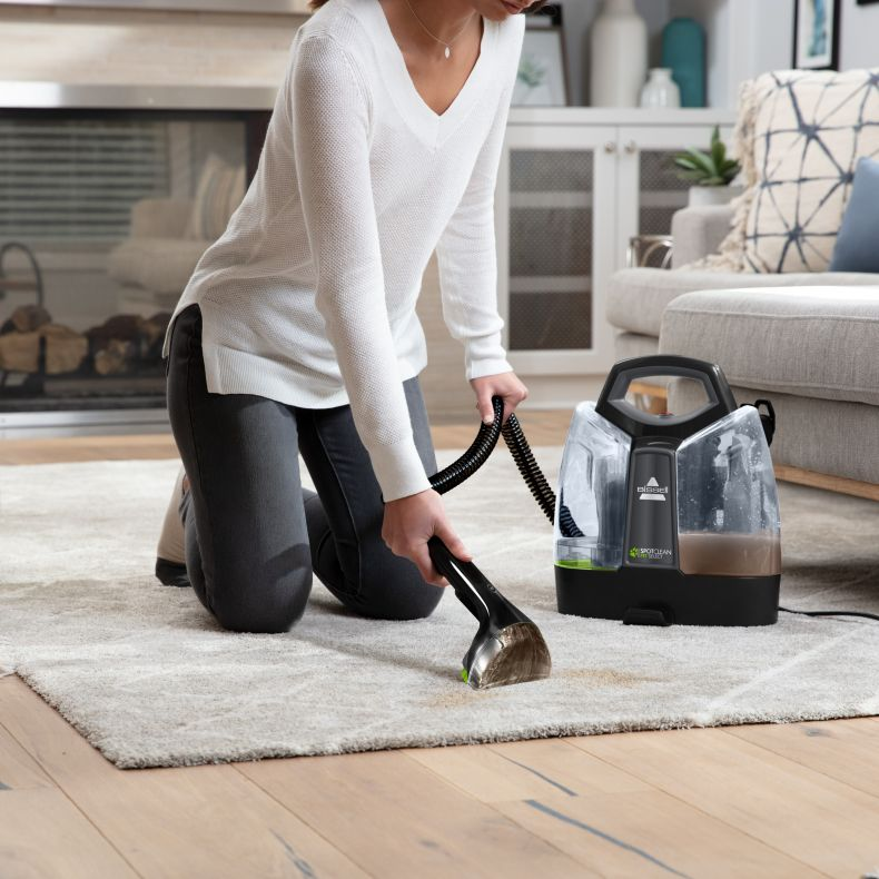 Una donna pulisce un tappeto con una macchina Bissell SpotClean. Il tappeto mostra le macchie in fase di pulizia.