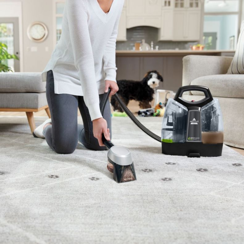 Donna che pulisce il tappeto con un pulitore Bissell. Cane che guarda sullo sfondo. Tappeto grigio con impronte di zampe.