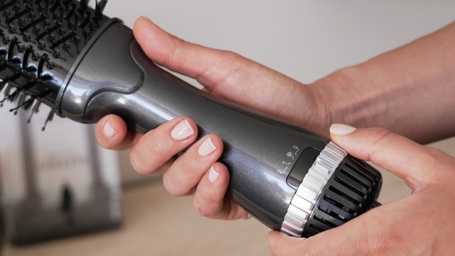 Hands holding a hair styling brush with a dial for heat settings.