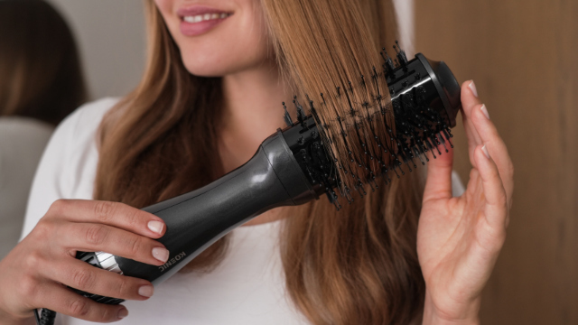 Woman styling hair with a black hair dryer brush. She is in front of a mirror.