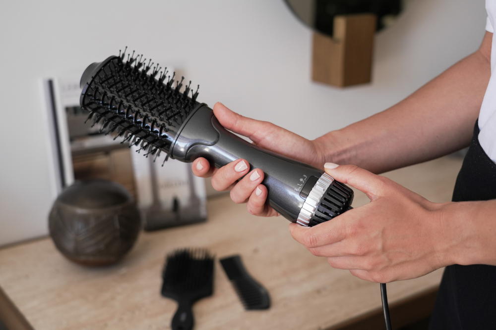 A person holds a gray hair dryer brush. Other hair tools are on the table. White wall in the background.