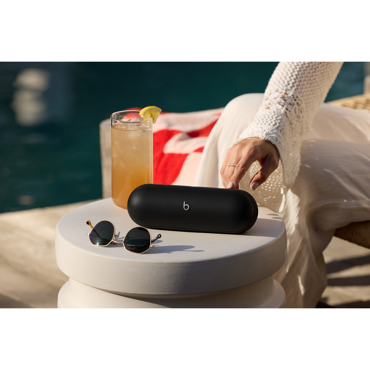 Altoparlante Beats nero e occhiali da sole su un tavolo bianco, con una bevanda e piscina sullo sfondo.