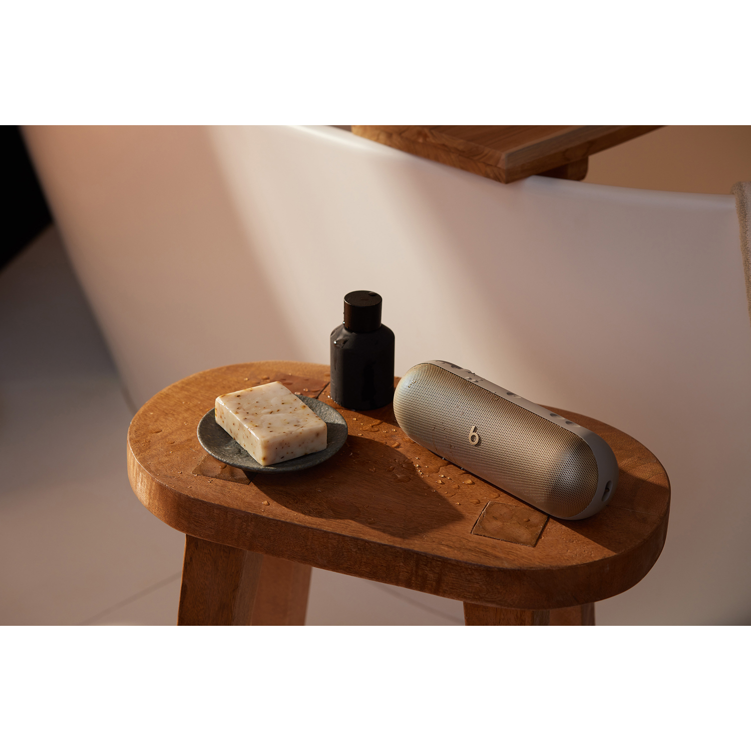Uno sgabello di legno con un portasapone, sapone e un altoparlante Beats accanto a una vasca da bagno.