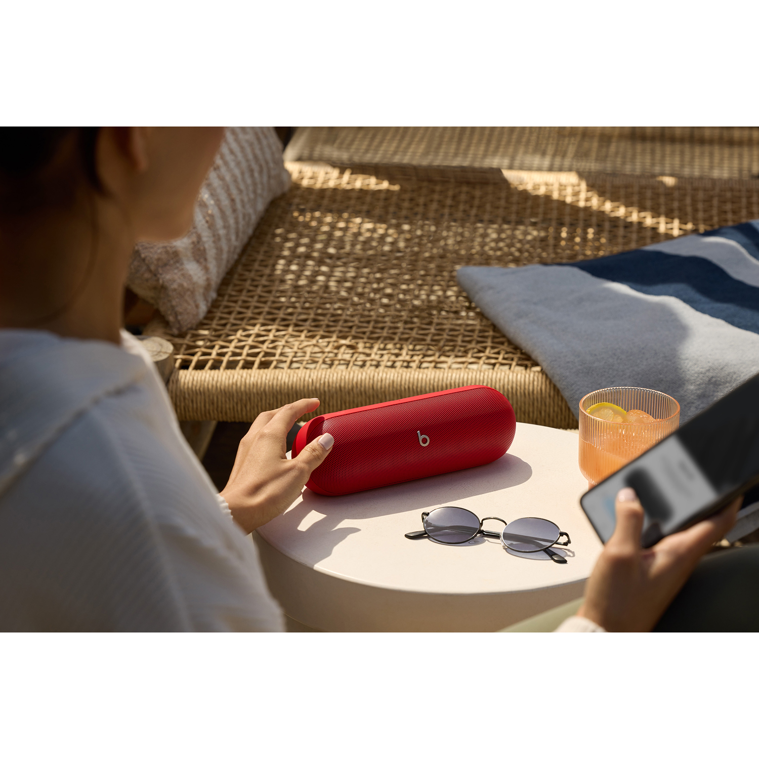 Donna che tiene in mano un altoparlante rosso. Occhiali da sole e un telefono sul tavolo. Dietro, mobili da spiaggia.