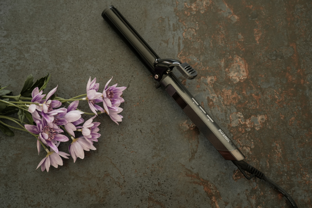A black curling iron next to a bouquet of purple flowers on a textured, dark surface.