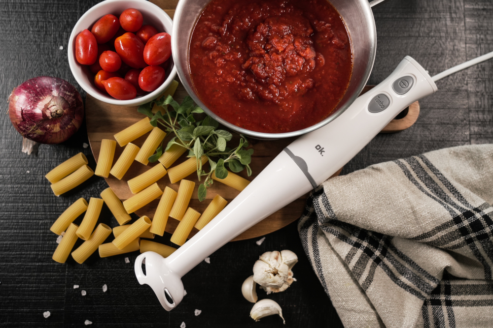 Overhead shot of pasta, tomatoes, sauce, and a blender on a wooden board and a dark surface.