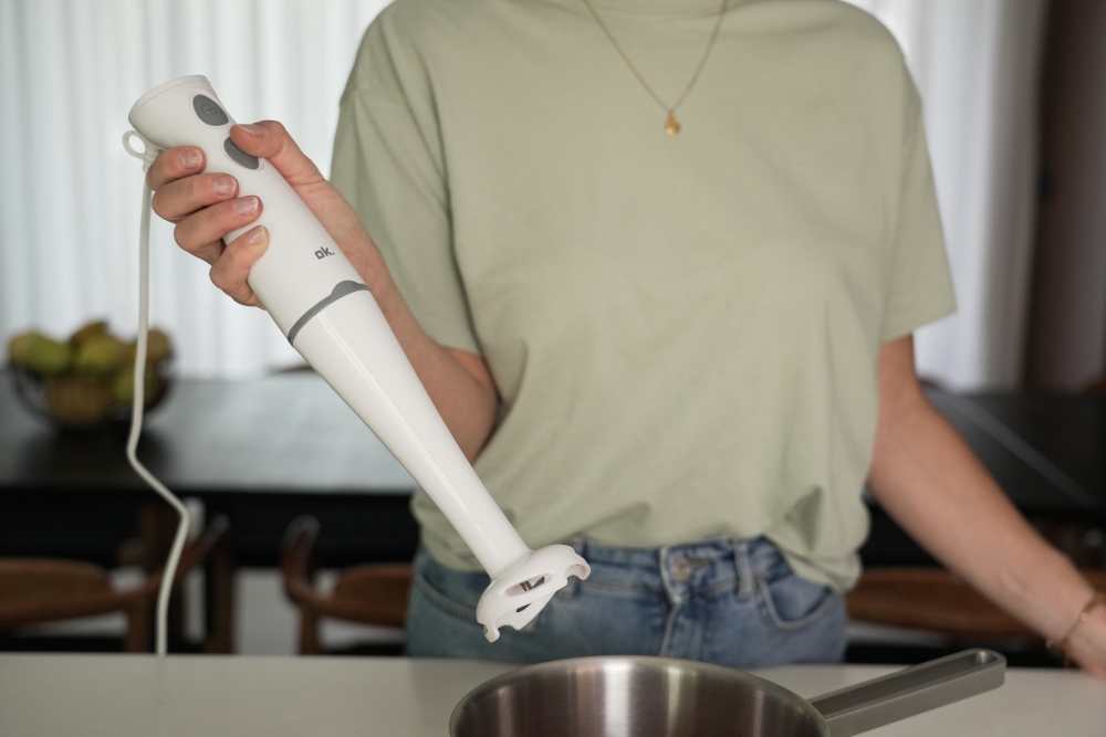 A person holds a white immersion blender over a stainless steel pot on a table.