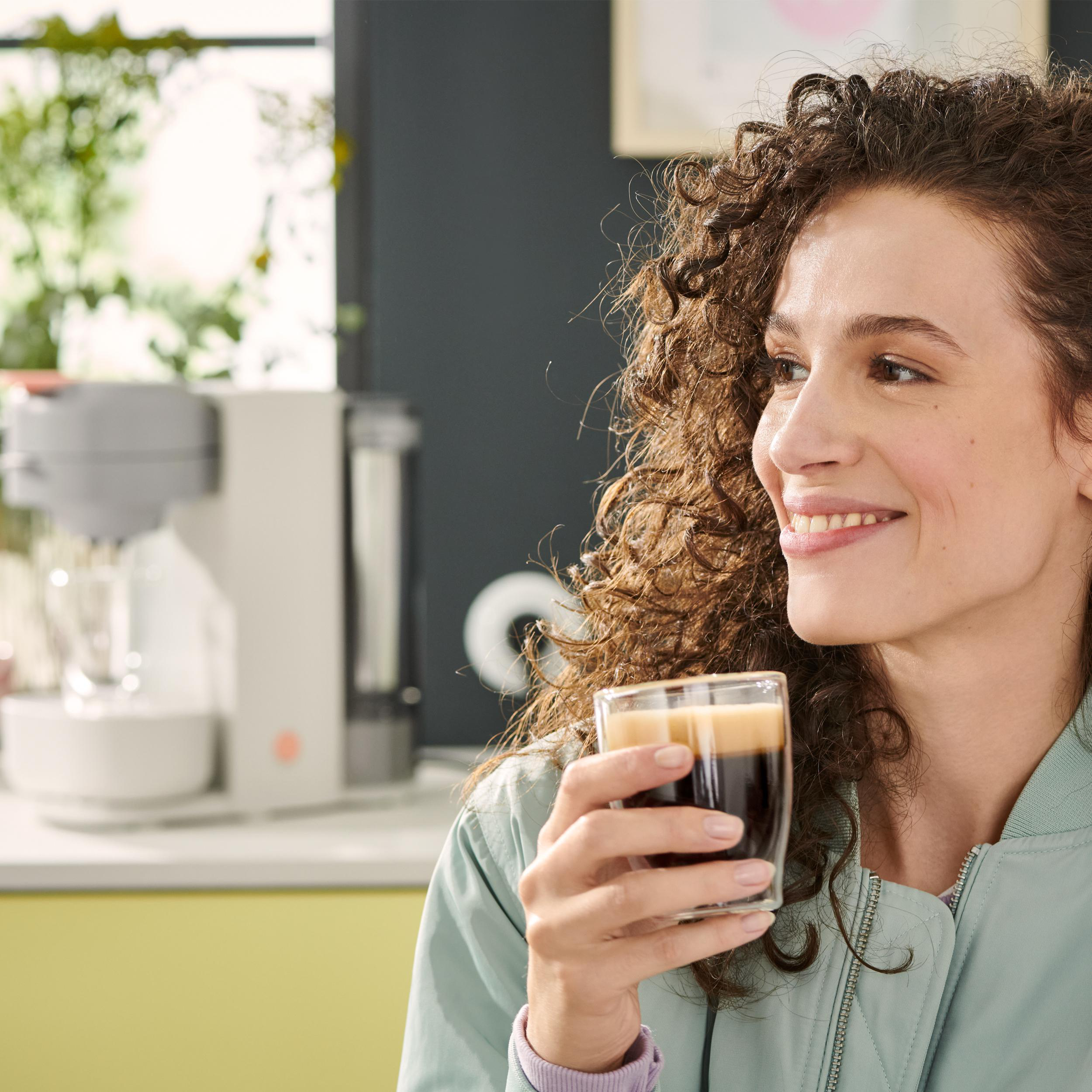 Lächelnde Frau, hält ein Glas dunklen Kaffee. Küchenhintergrund mit Kaffeemaschine.
