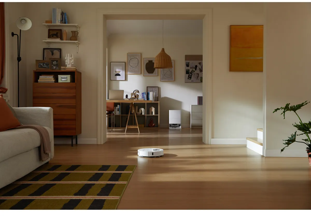 Un aspirateur robot sur un plancher en bois. En arrière-plan, il y a un salon avec un canapé et des œuvres d'art.