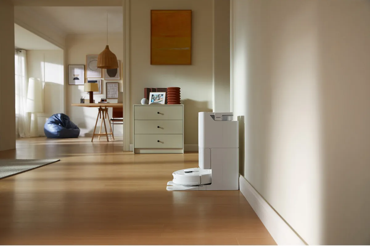 Intérieur d'une maison moderne avec un aspirateur robot au sol. Un couloir mène à un salon.