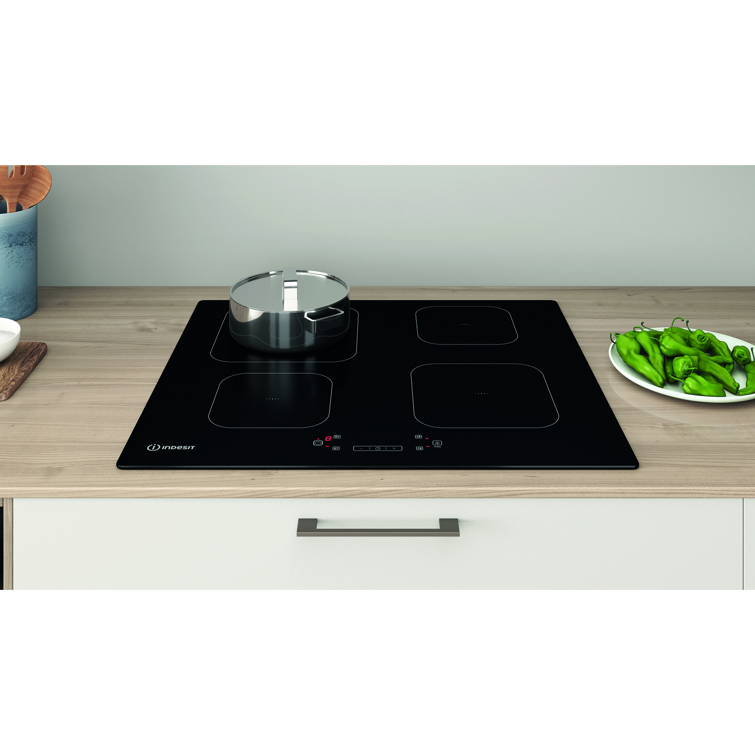 Piano cottura Indesit nero con pentola, quattro zone cottura e piatto di peperoni verdi.
