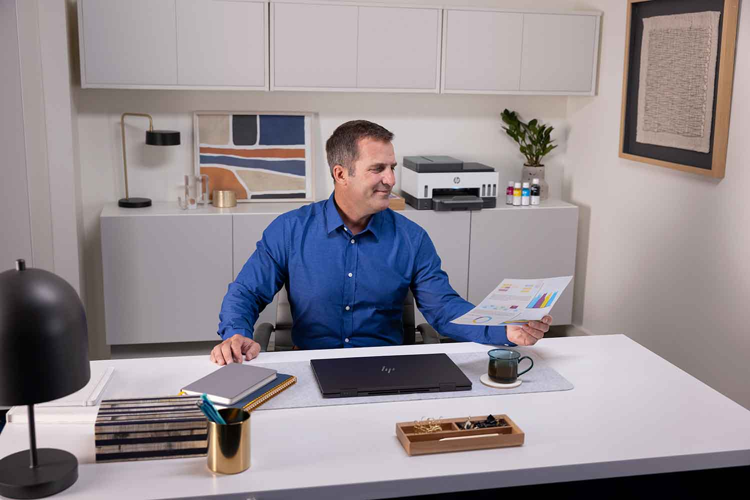 A man sits at a white desk, looking at a document. There's a printer and office supplies nearby.