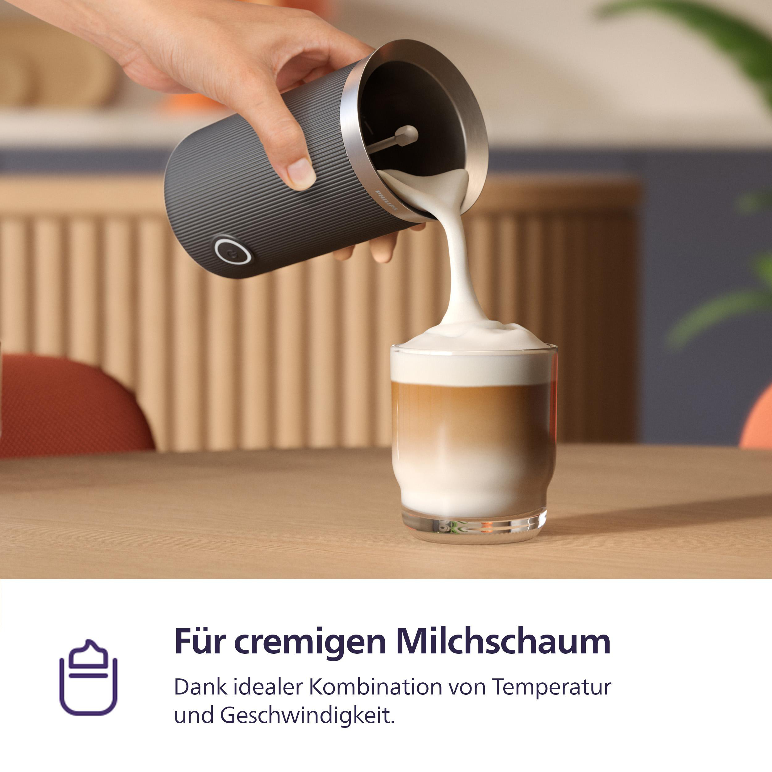 Eine Hand gießt Milchschaum aus einem Aufschäumer in ein Glas geschichteten Kaffees. Der Tisch ist aus Holz.