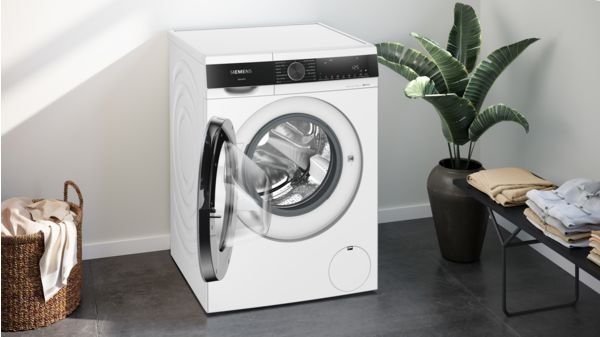 Una lavadora blanca con la puerta abierta, junto a una cesta de ropa y plantas.