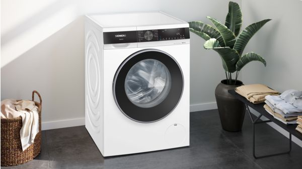 Lavadora blanca con puerta y panel de control negros, junto a una planta y una cesta de lavandería.