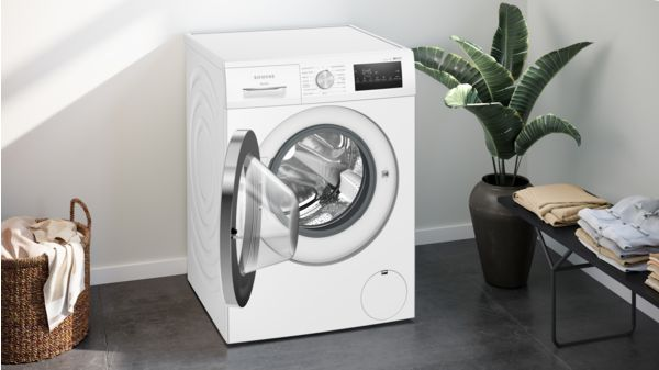 Lavadora blanca con puerta abierta. Cesta de ropa y planta al fondo.