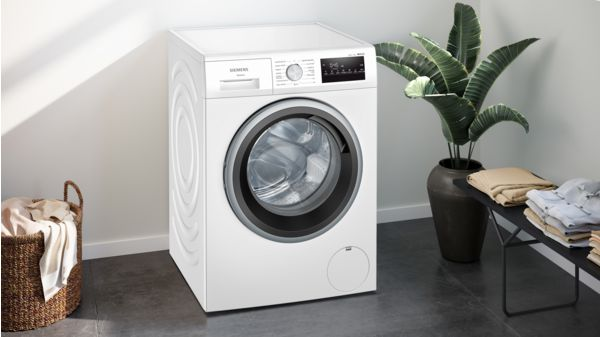 Lavadora Siemens blanca junto a una planta y una cesta de ropa.