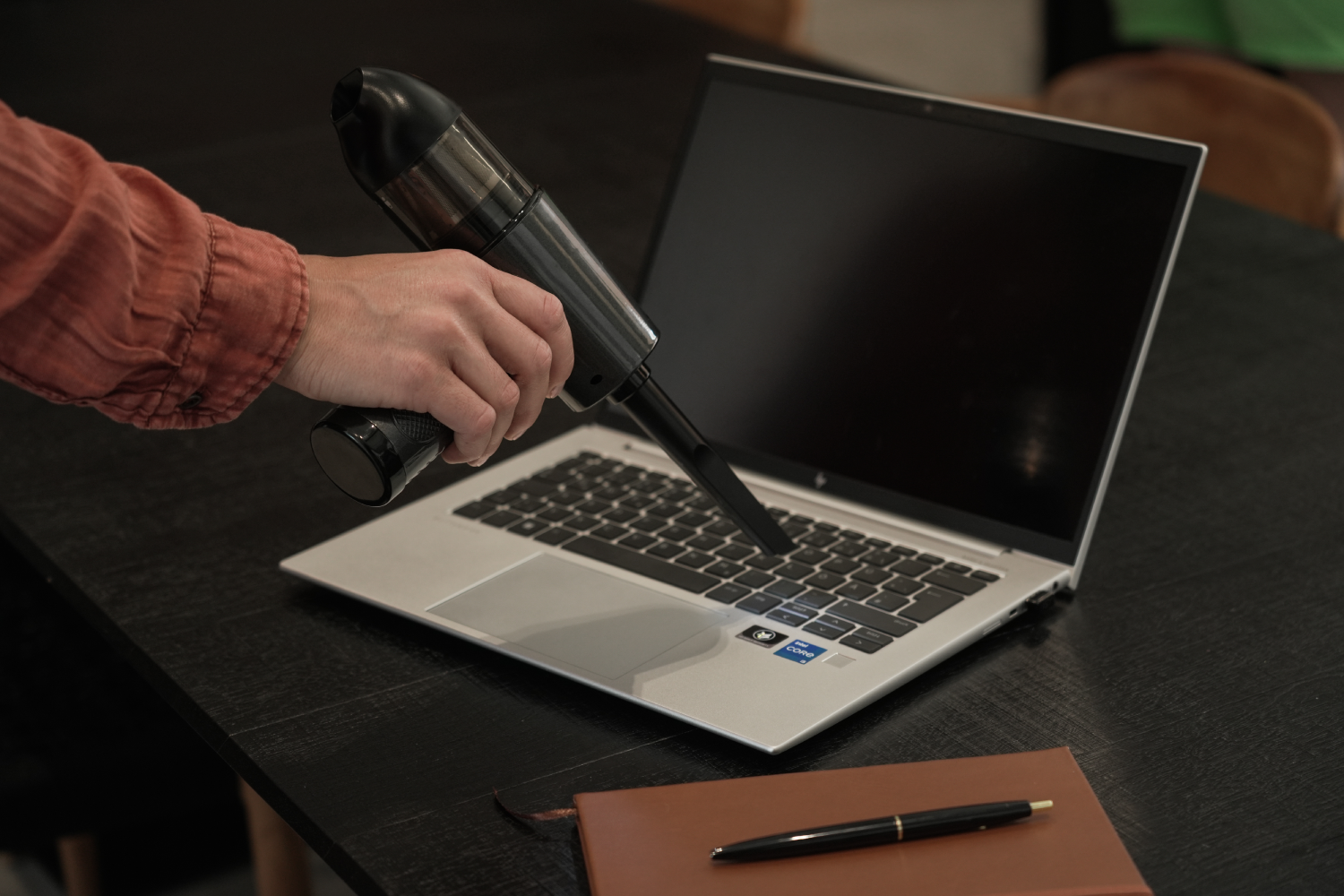 Persona limpiando el teclado de un portátil con una aspiradora de mano. Portátil, bolígrafo y bloc de notas en la mesa.