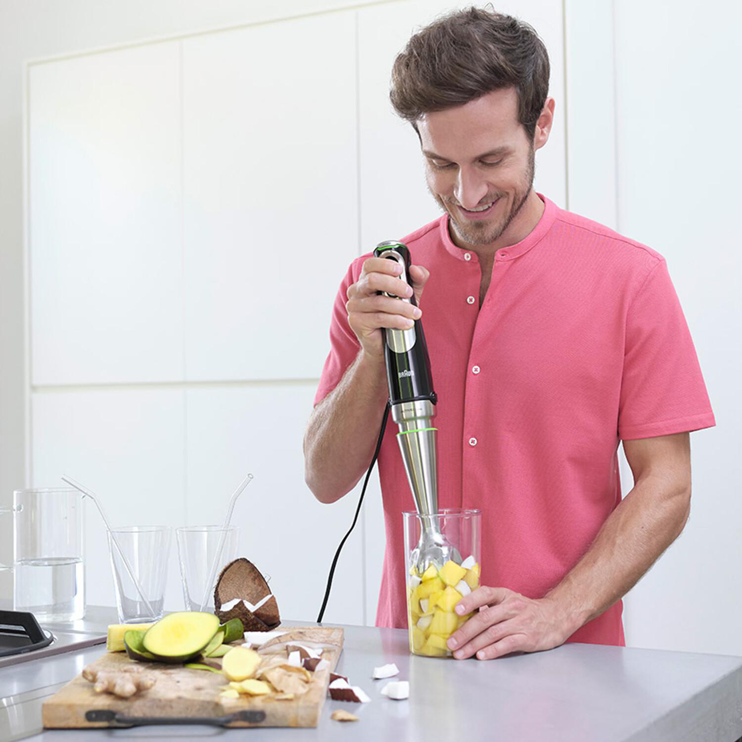 Mann, der einen Stabmixer verwendet, um Zutaten in einem Glas zu mischen, mit Zutaten auf einem Holzbrett.