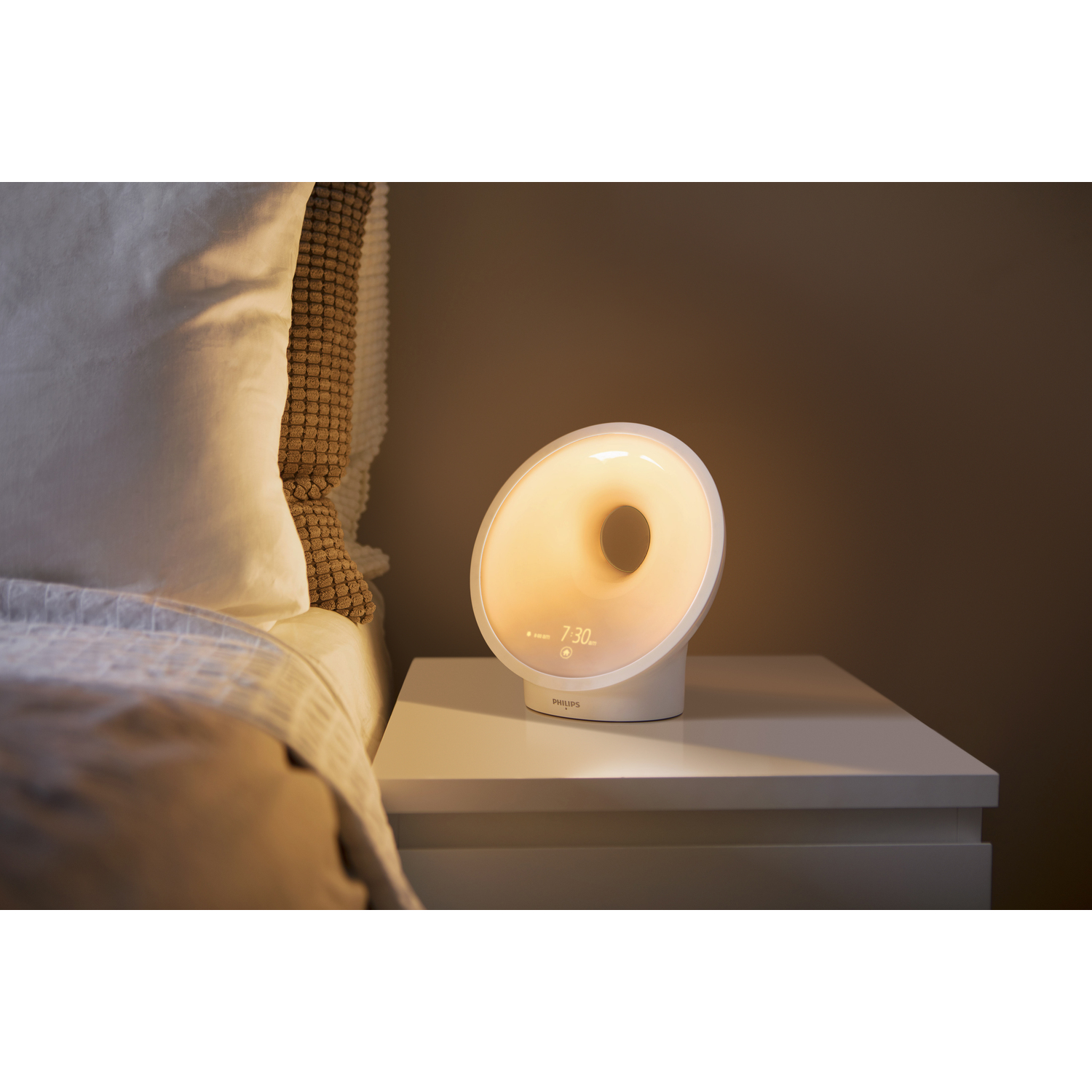 Un réveil blanc en forme d'anneau est posé sur une table de chevet à côté d'un lit.
