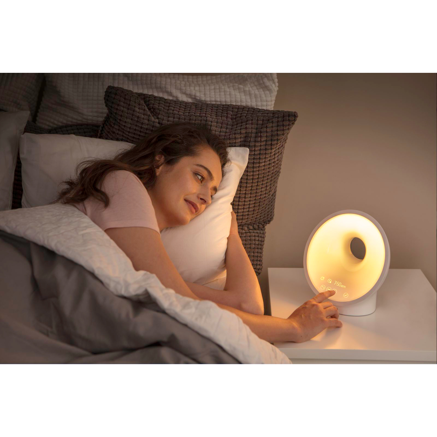 Une femme est allongée dans son lit, tendant la main vers un réveil blanc. La lumière brille de l'horloge vers la table de chevet.