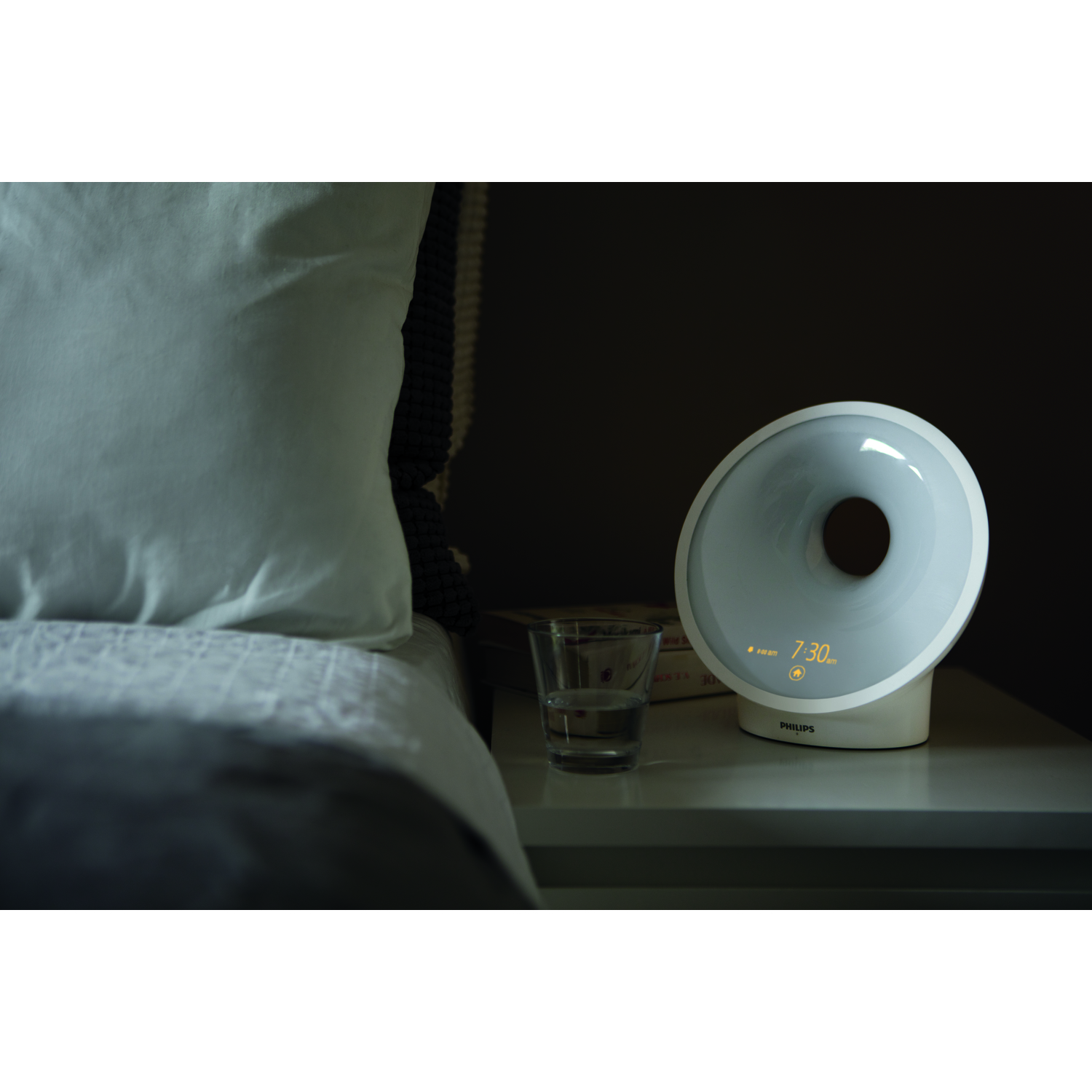 Réveil blanc avec écran rond affichant 7h30, à côté d'un verre d'eau et d'un livre.
