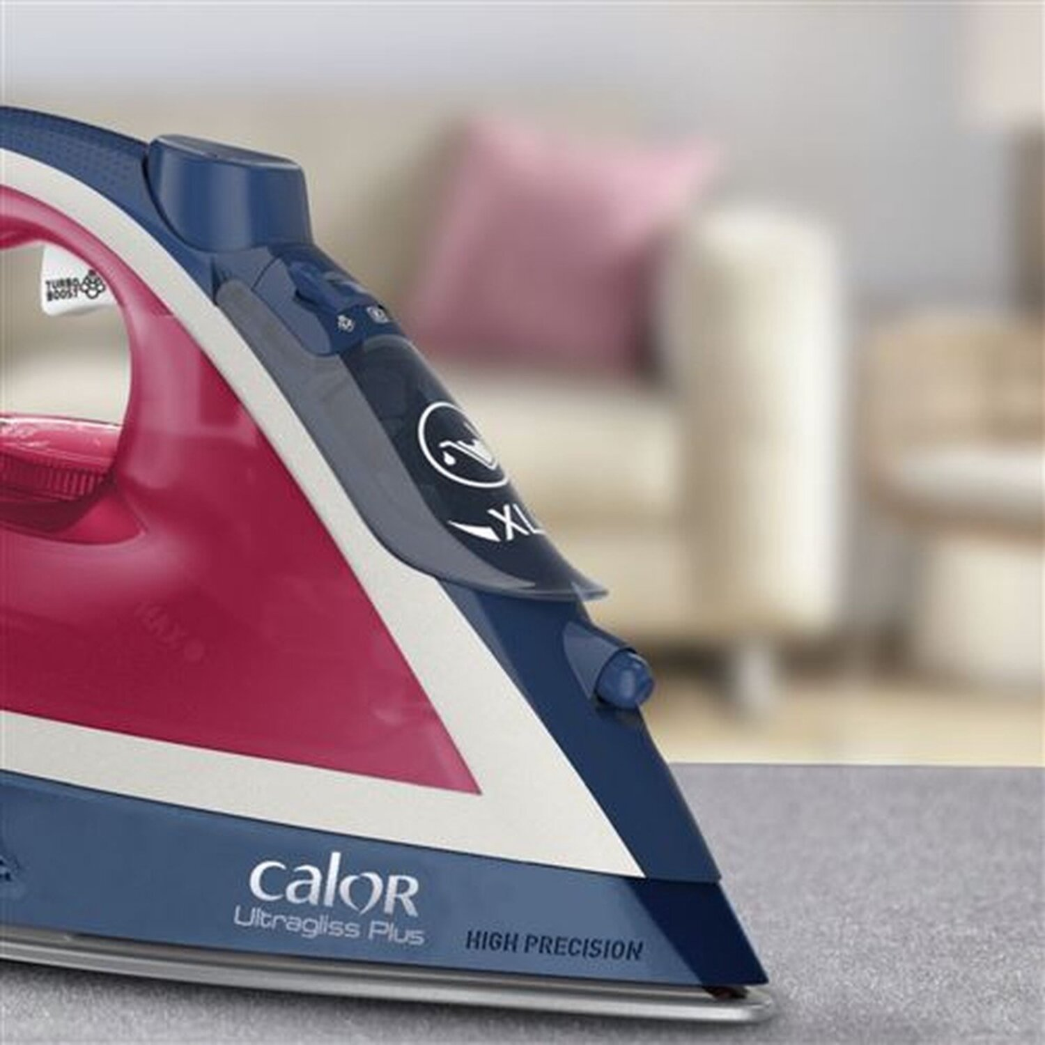 Gros plan d'un fer à repasser rouge et bleu sur une surface grise, avec un arrière-plan flou de salon.
