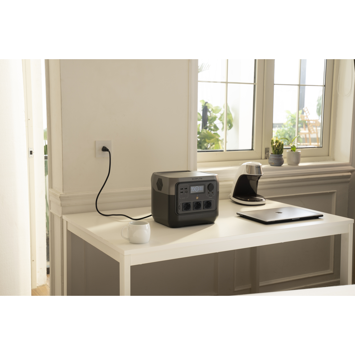 Une station d'alimentation portable sur une table blanche, branchée au mur. Un ordinateur portable, une cafetière et une tasse sont sur la table.