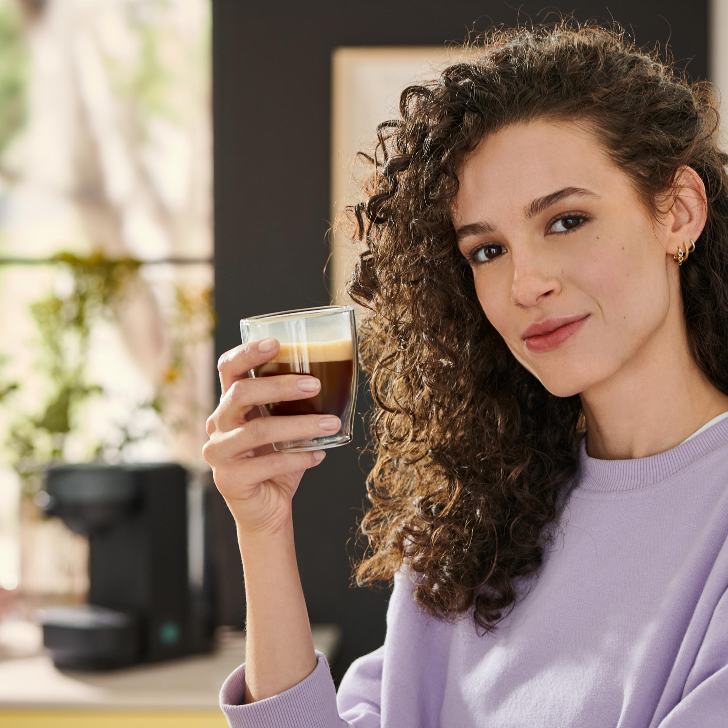 Eine Frau mit lockigem Haar hält ein Glas Kaffee, im Hintergrund eine schwarze Kaffeemaschine.