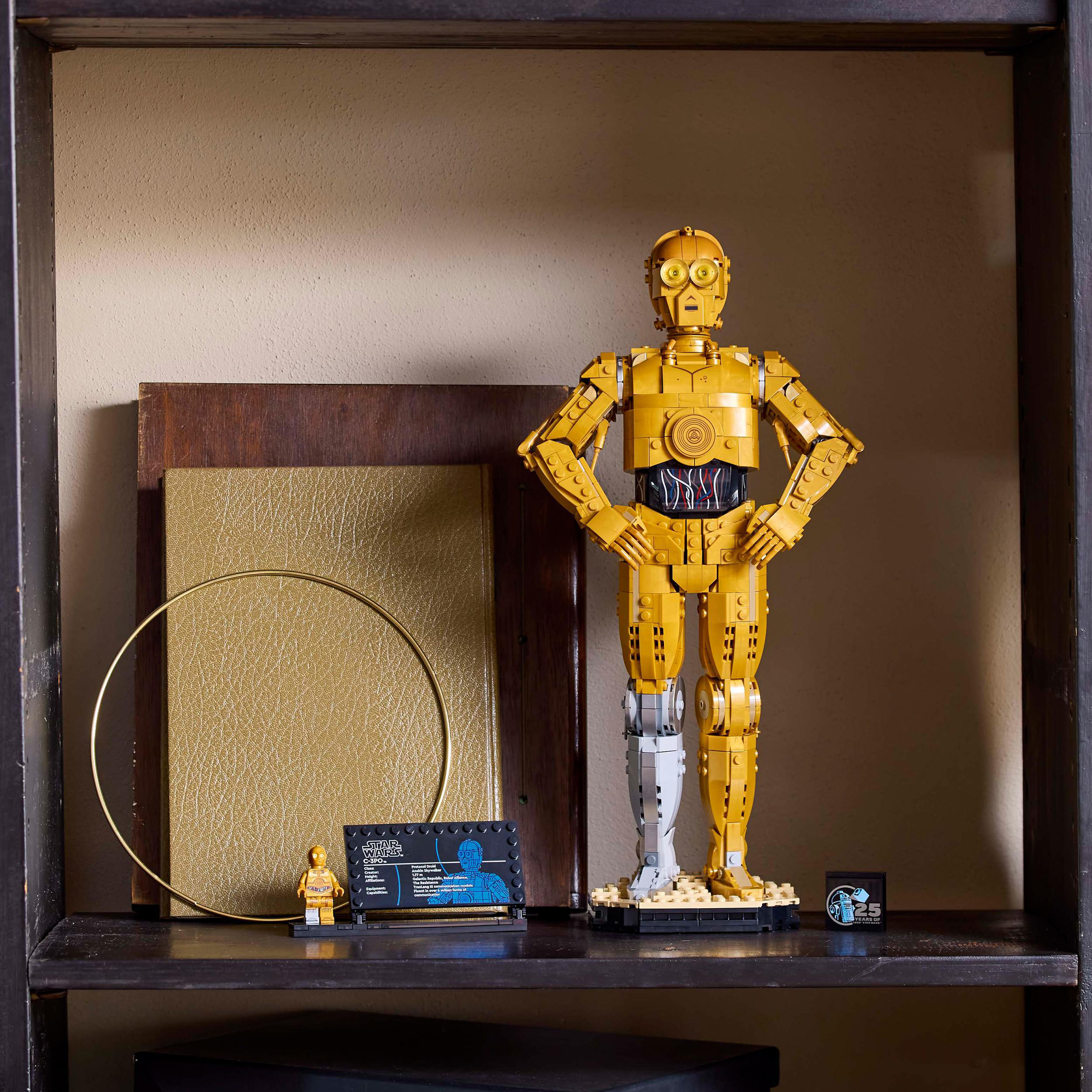 LEGO C-3PO Figur auf einem Regal mit anderen Gegenständen. Eine kleine LEGO Figur und eine blaue Plakette liegen daneben.