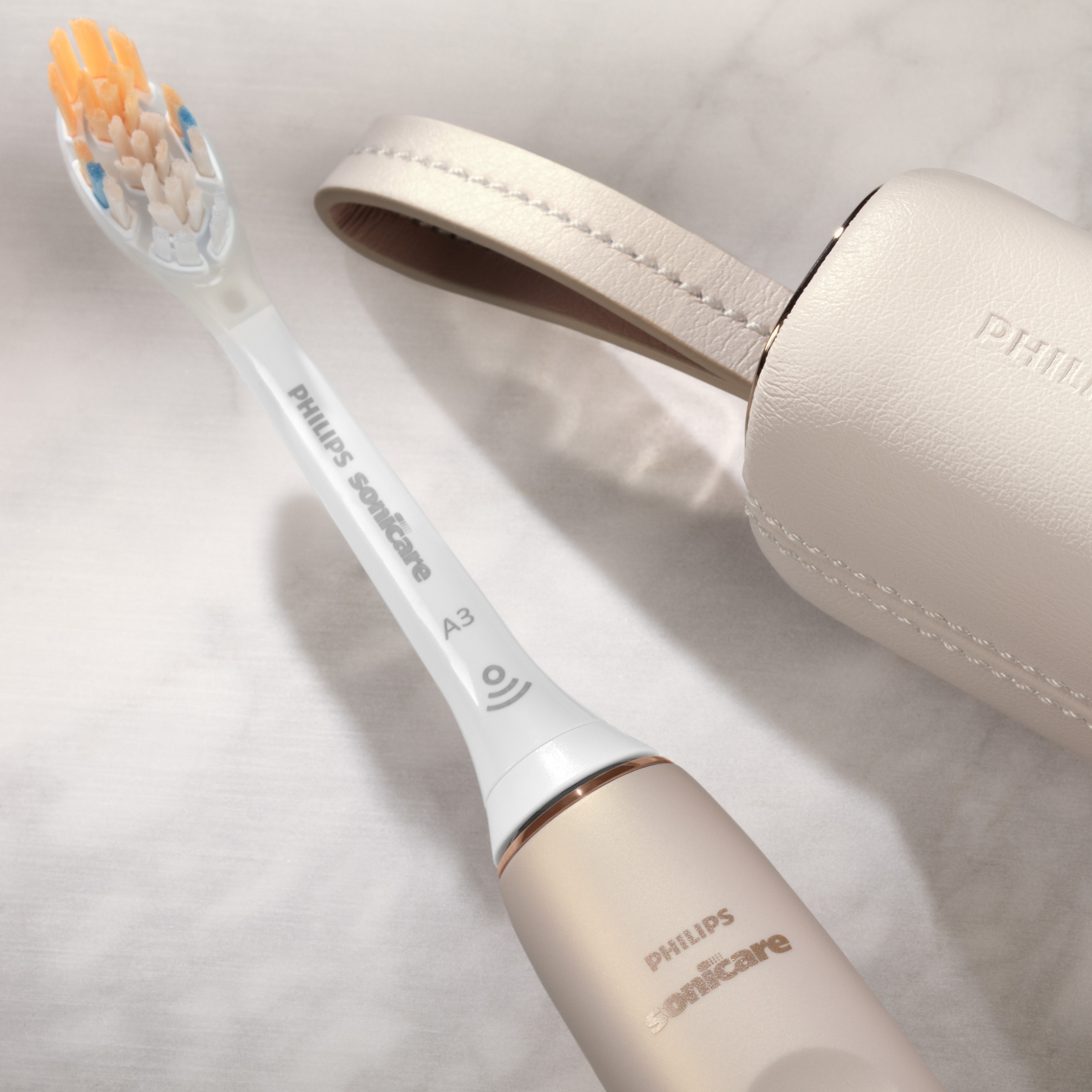 Elektryczna szczoteczka do zębów i etui. Szczoteczka jest biała i złota, z pomarańczowym włosiem. Etui jest beżowe.