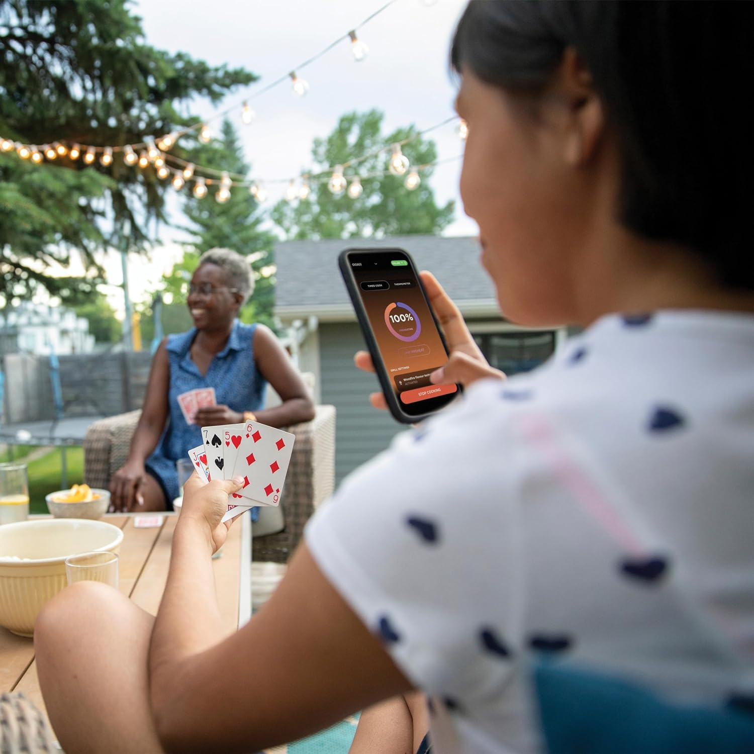 Mujer usando teléfono con app, sosteniendo cartas, otra mujer en el fondo jugando cartas, al aire libre.