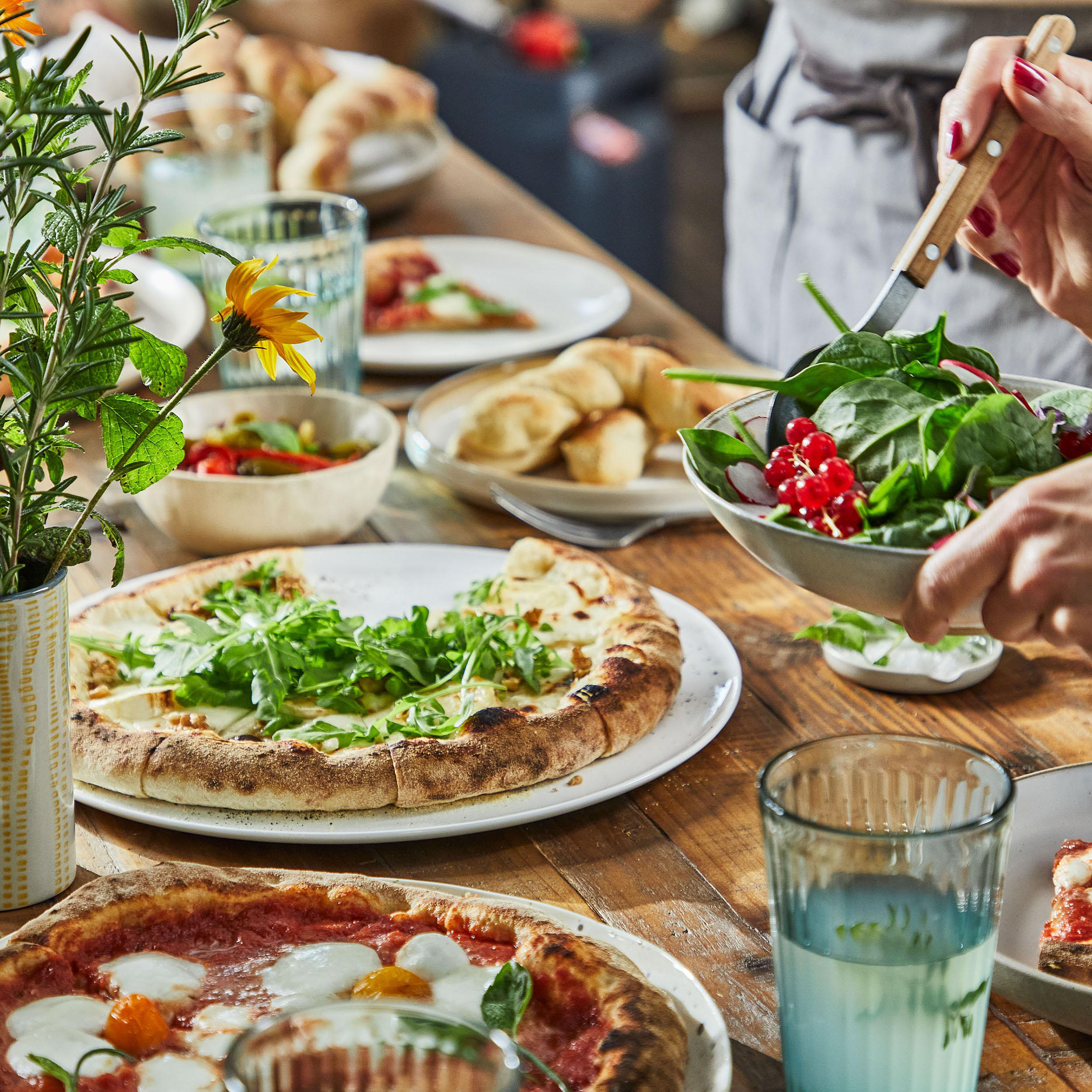 Ein Tisch mit Essen: Pizza, Salat, Croissants und Getränke. Person hält Salat.