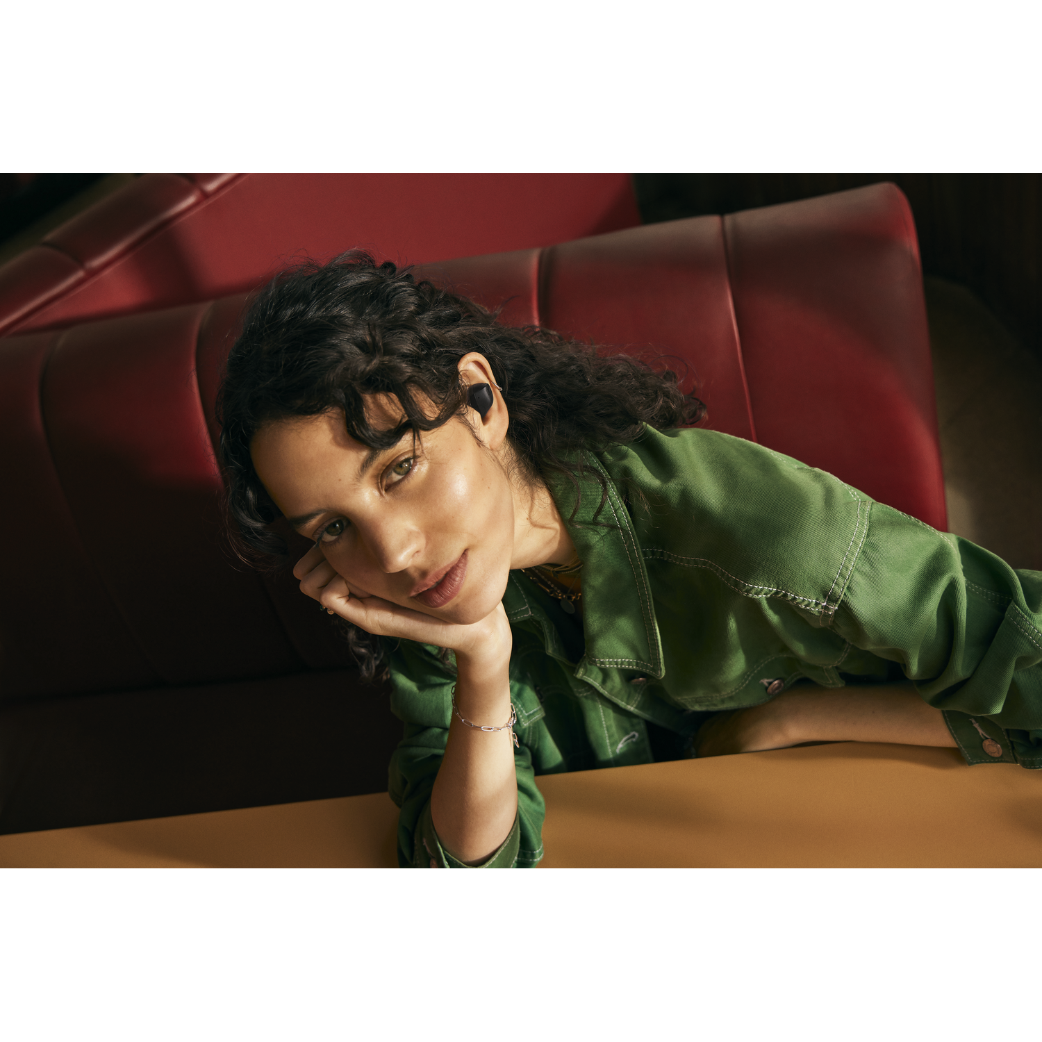 Young woman with earbuds, green jacket, leaning on a table.