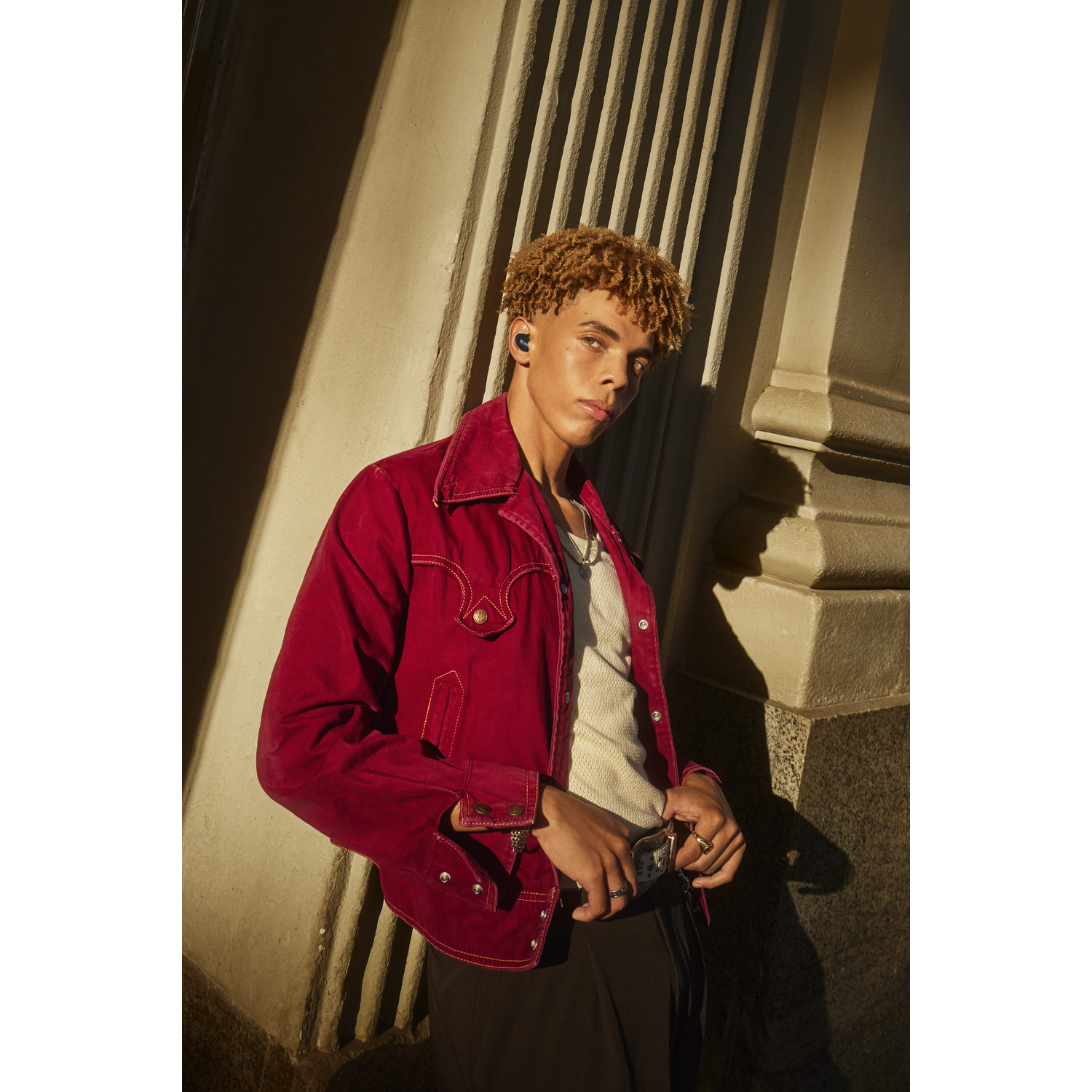Young man in red jacket and black pants, wearing earbuds and leaning against a wall.