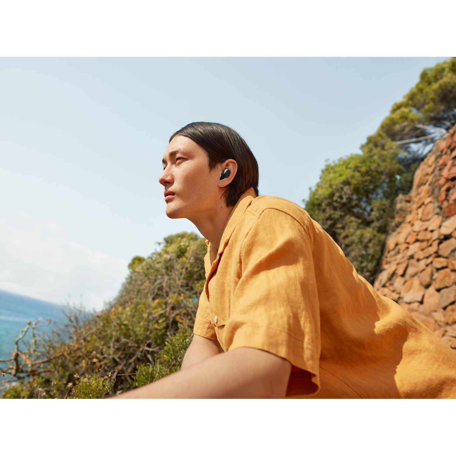 A man with headphones looking away, in a sunny environment with green trees and blue sky.