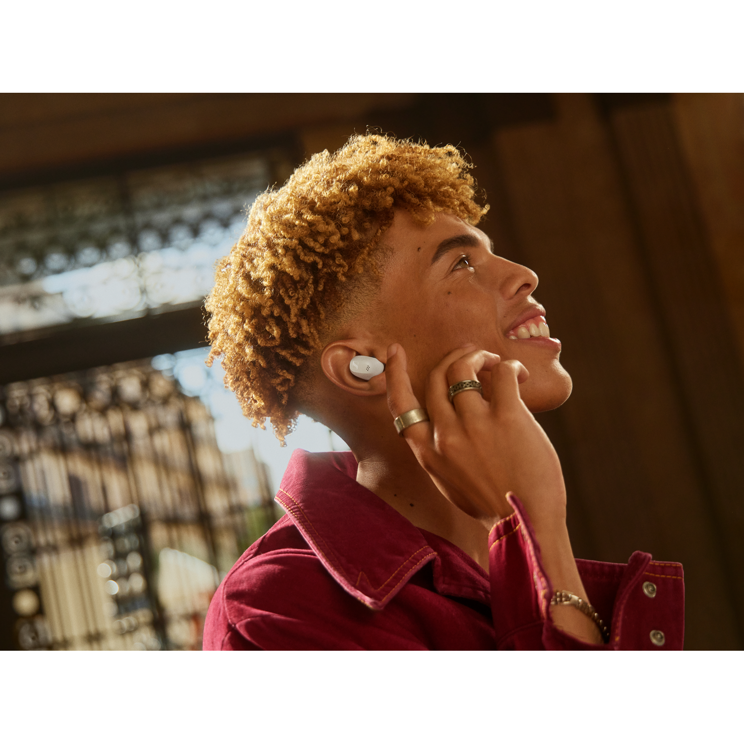 A young man with curly hair smiles, wearing an earbud, looking up.